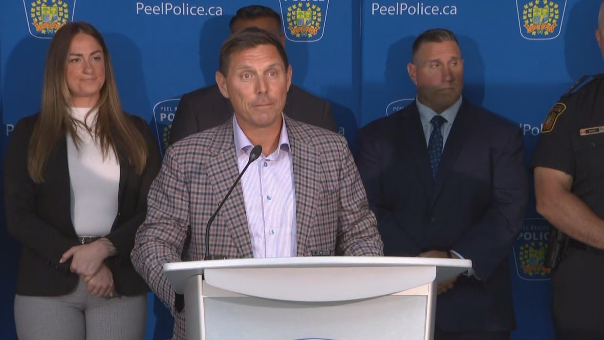 Man wearing blazer and button-up stands in behind white podium, with others dressed up nicely behind him