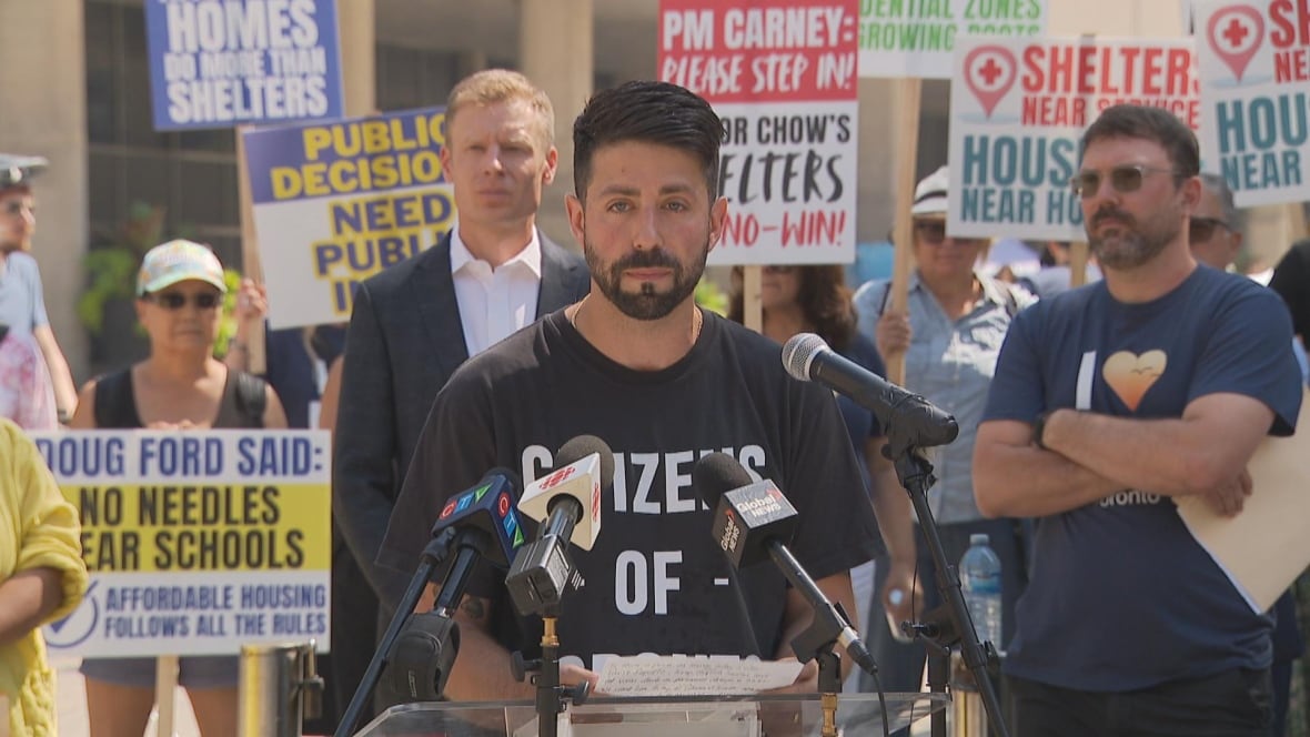 man wearing black shirt, speaking on a mic, with signs behind him.