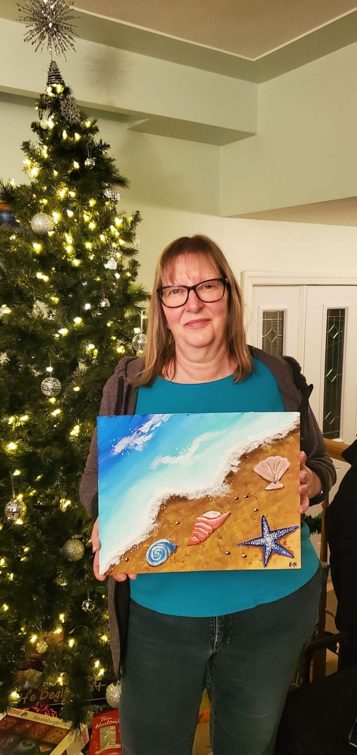 Person looking at the camera, standing in front of a decorated tree, while holding a painting.