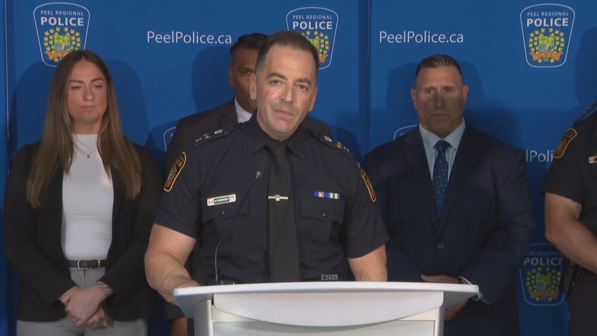 Man in cop uniform stands behind white podium, with others nicely dressed behind him