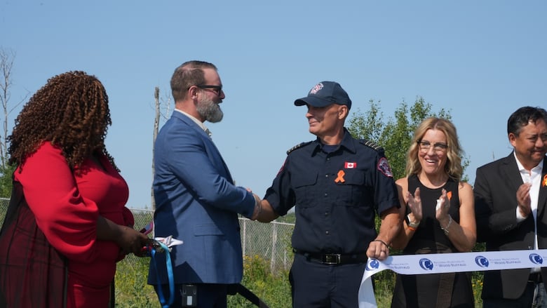 Bearded man in a suit shakes hands with a thin clean-shaven man in an emergency services uniform. Several people clap beside them.