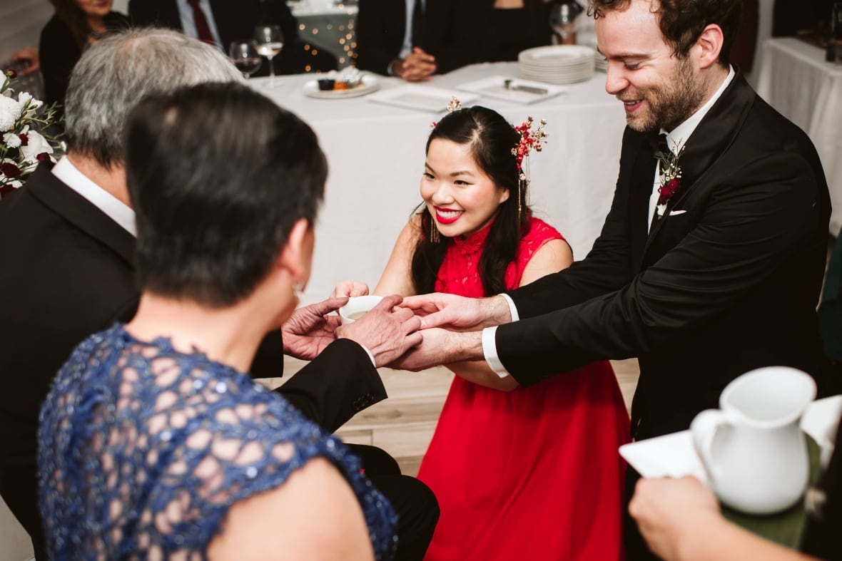 A smiling man and a smiling woman kneel on the floor and hand a teacup to a seated older couple.  
