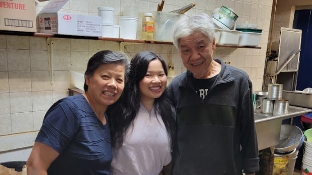 Two smiling women and a smiling man pose for a photo inside a large kitchen.  