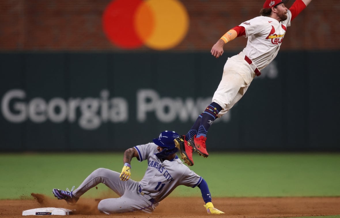 A player in a grey uniform slides into a base as a player in a white uniform leaps above him to try to catch a ball