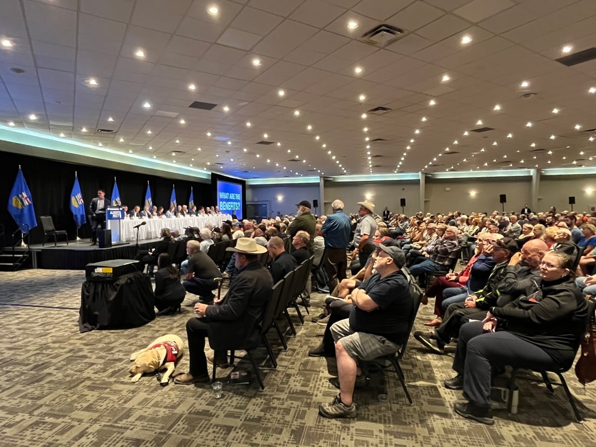 A panel of people, including Premier Danielle Smith, is visible on the left sitting at a long table draped in white cloths. A crowd of rows of people seated and looking at the panel is assembled in a ballroom with overhead pot lighting.