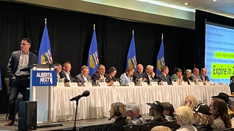 A panel of sixteen people, including Alberta premier Danielle Smith, sits at a long white table on a stage. Six Alberta flags hang on poles in the background.