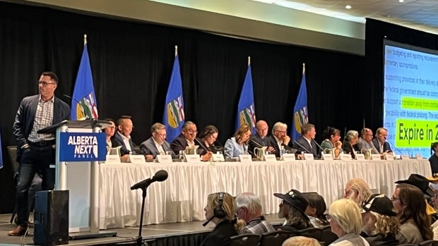 A panel of sixteen people, including Alberta premier Danielle Smith, sits at a long white table on a stage. Six Alberta flags hang on poles in the background.