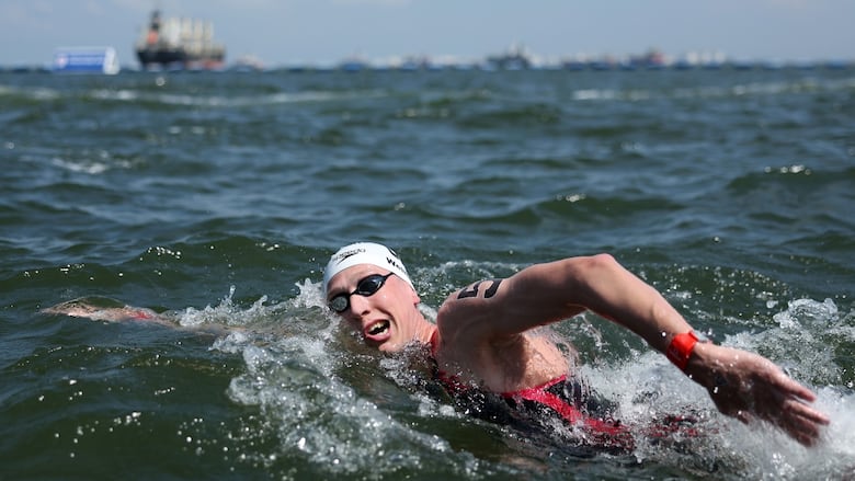 A men's swimmer with swim goggles and a swim cap on, is shown in action in an open body of water with a large boat in the far background.