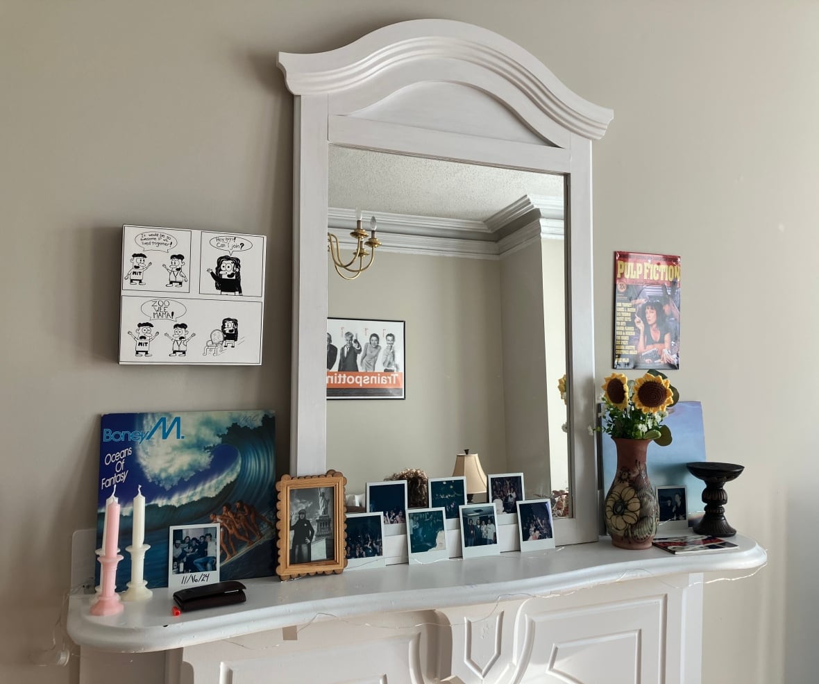 A fireplace mantle with a mirror and a row of photos propped up against it, among other items like records, candles, and a flowerpot.
