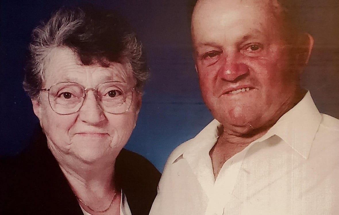 An older couple posing together, the woman wearing glasses and a brooch, and the man in a short-sleeved shirt and wristwatch