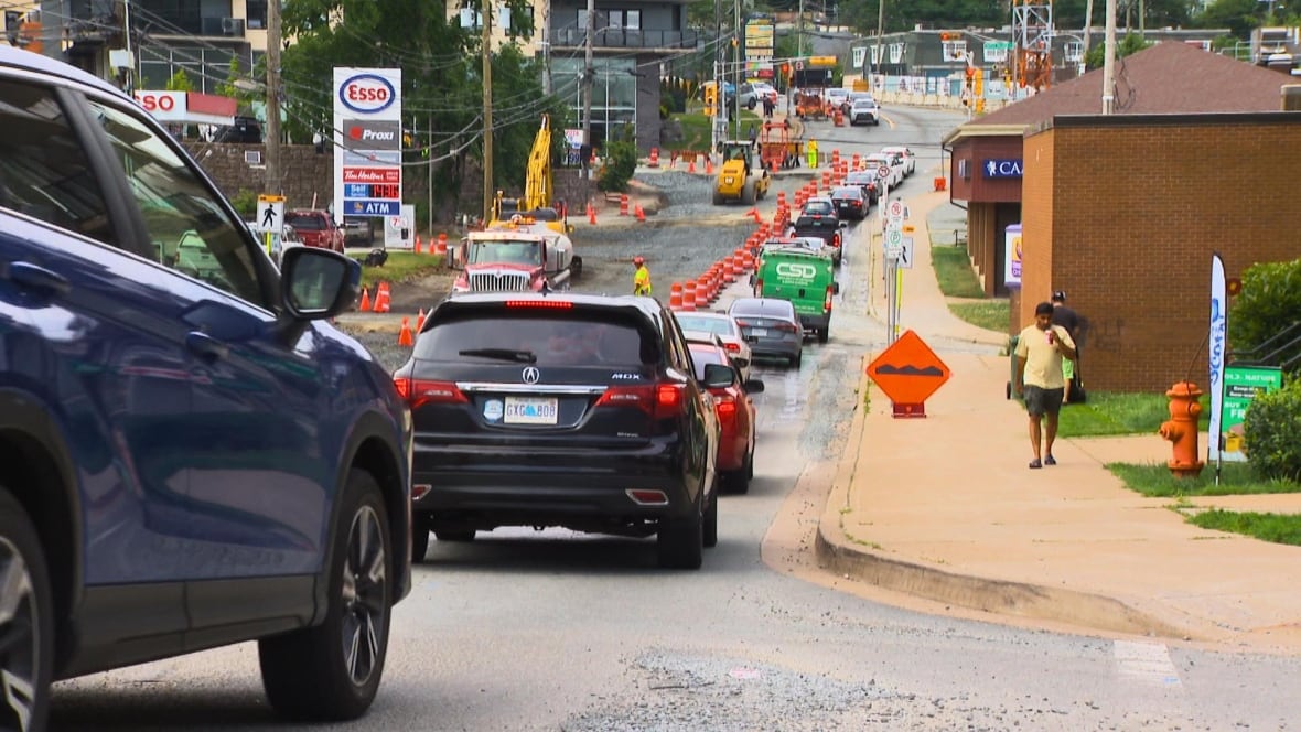Cars line up in busy street, full of cones, machinery, and unfinished streets. There are a few construction workers in bright yellow uniforms. 