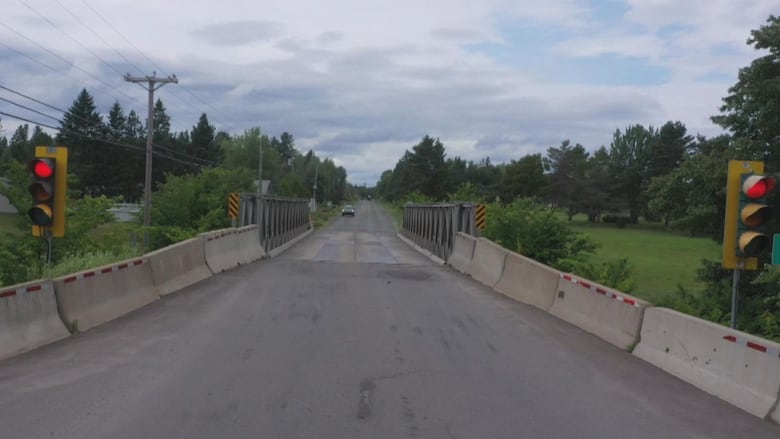 A shot of a bridge reduced to a single lane, bookended by traffic lights, spans a shallow river.