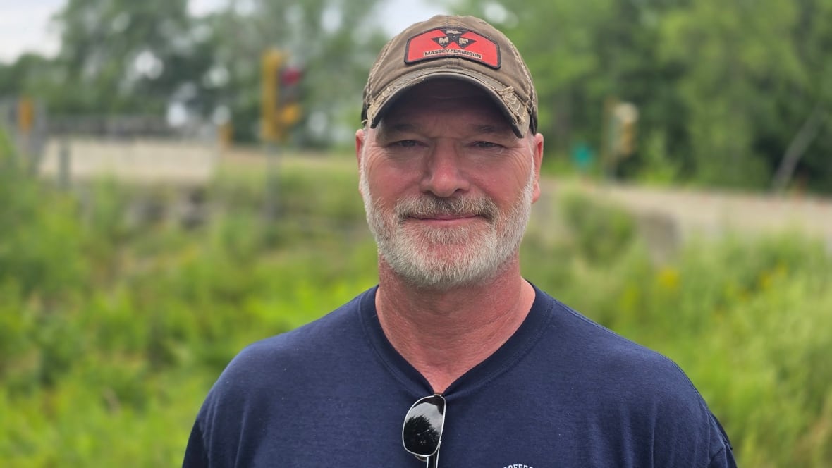 A man with a white beard, wearing a blue T-shirt and baseball cap smiles at the camera with a set of red lights and a bridge in the background.