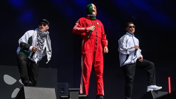 Mo Chara, DJ Provaí and Móglaí Bap of Kneecap perform on the West Holts stage during day four of Glastonbury festival 2025 at Worthy Farm, Pilton on June 28, 2025 in Glastonbury, England. Established by Michael Eavis in 1970, Glastonbury has grown into the UK's largest music festival, drawing over 200,000 fans to enjoy performances across more than 100 stages. In 2026, the festival will take a fallow year, a planned pause to allow the Worthy Farm site time to rest and recover. Three men standing on an outdoor concert stage. From left to right: A man wearing a white jacket, black pants, a black and white keffiyeh scarf around his neck, a black ballcap and sunglasses; a man in a red jumpsuit stands centre stage with a green, white and green Irish flag ski mask over his head and face; another man wearing a white jacket, with black stripes on the sleeves, black pants and sunglasses.