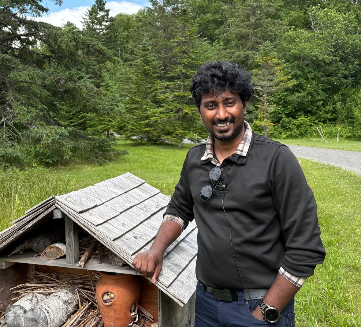 Man in a black shirt stands in front of a "bee hotel" in a public garden.