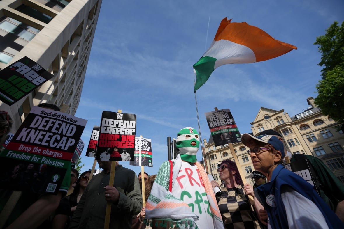 People holding protest signs outside with a large green, white and orange Irish flag waving in the wind above. 