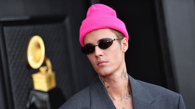A man in a pink toque and sunglasses with neck and chest tattoos, wearing a grey suit jacket.