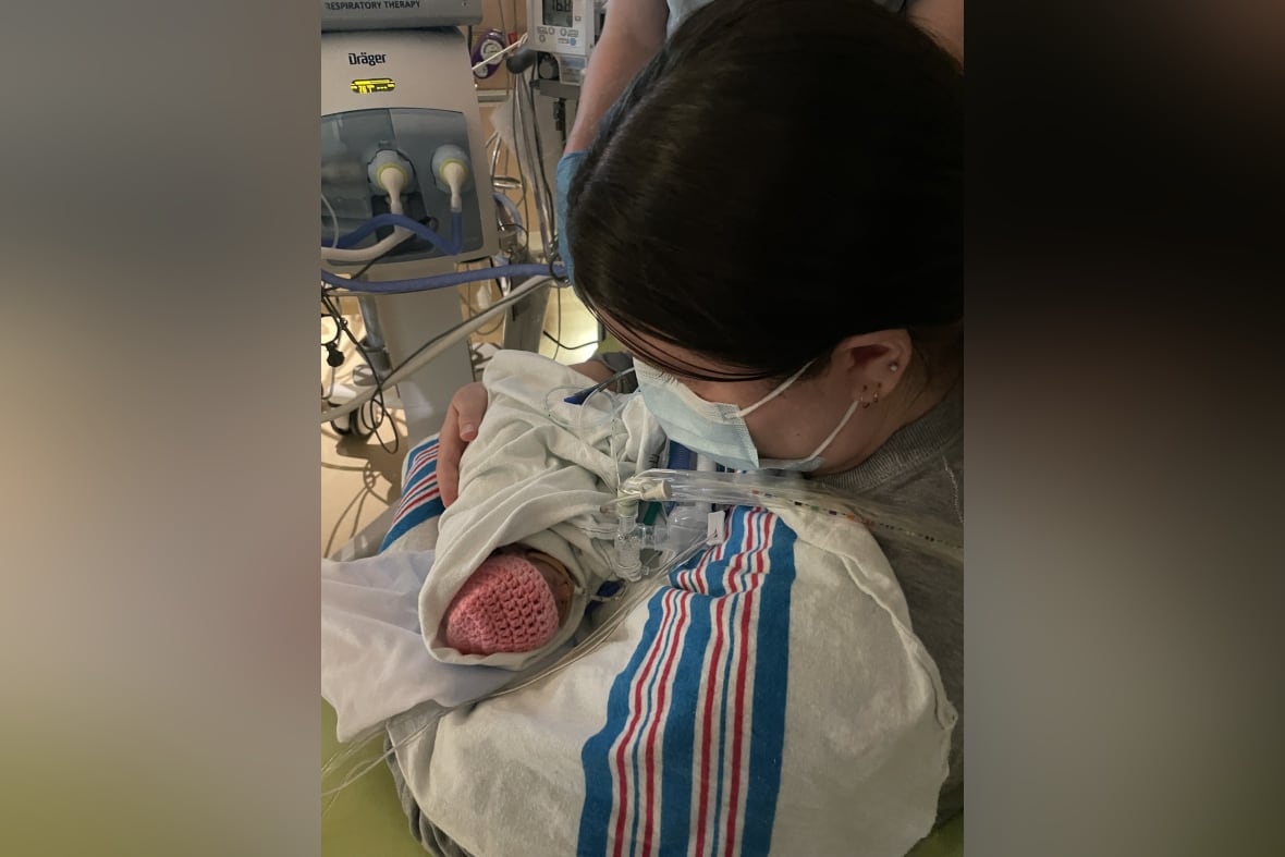 A dark-haired woman in a mask holds a tiny swaddled baby with a pink cap.
