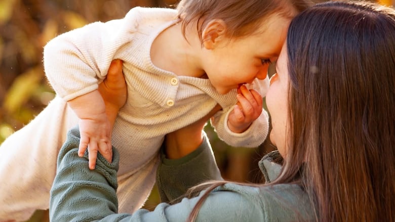 A dark haired woman in a blue coat holds a baby up against the background of trees.