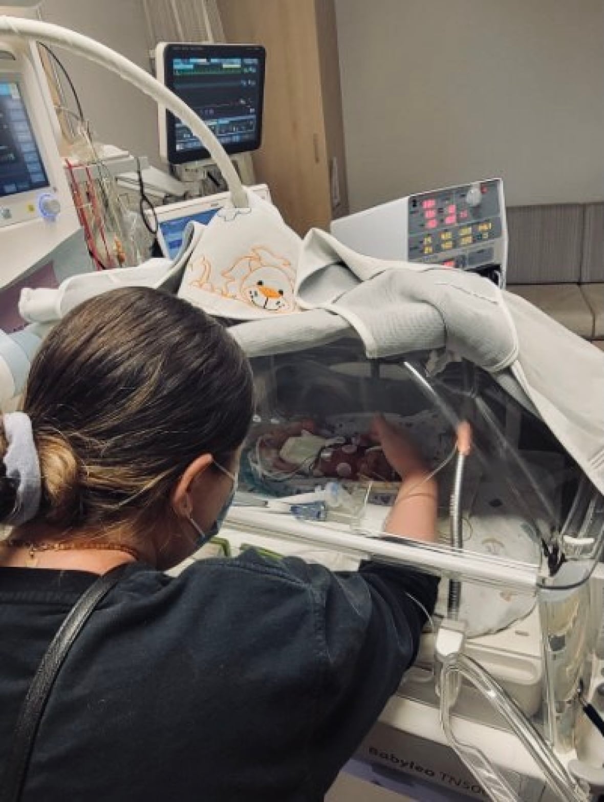 A woman in a black shirt puts her hand into an incubator, in which lies a baby hooked up to monitors.