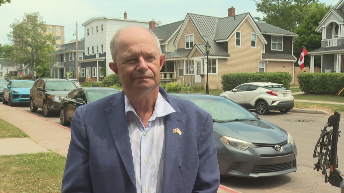 A man in a blue blazer stands on a residential street with parked cars and houses behind him.