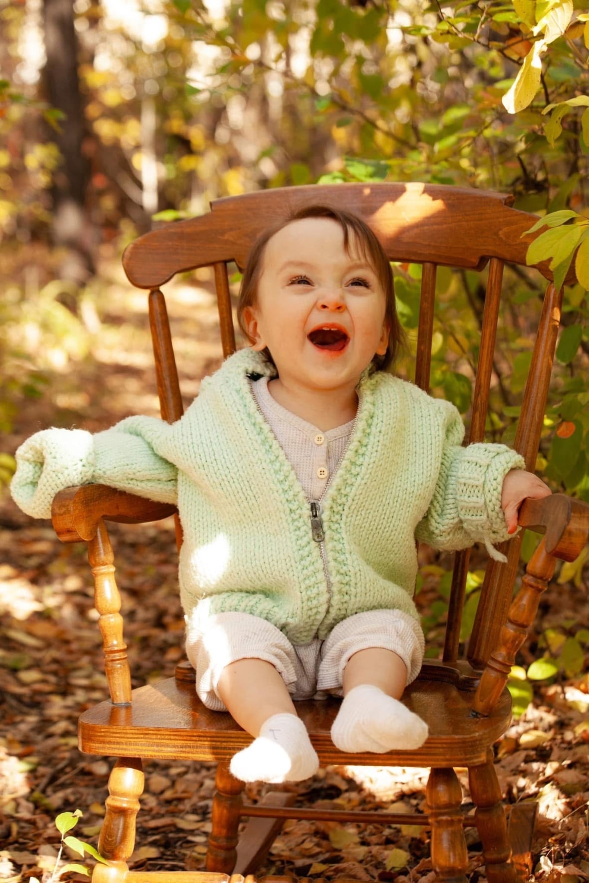 A little girl in a green cardigan smiles as she sits on a wooden chair.