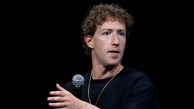 A curly haired cleanshaven man gestures while speaking and seated.