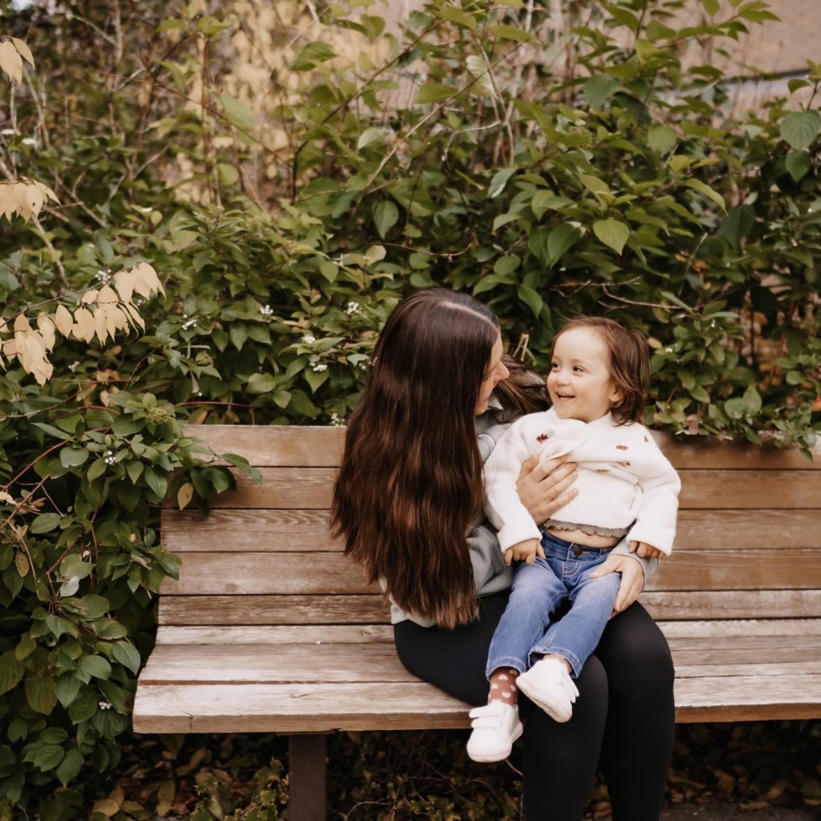 A dark-haired woman smiles at the child sitting on her lap, as she sits on a wooden bench surrounded by plants.