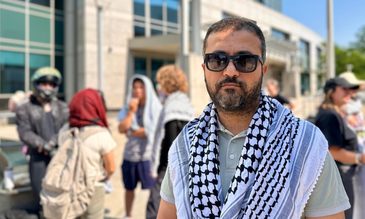 A person wearing a keffiyeh and sunglasses stands before a group outside a city building.