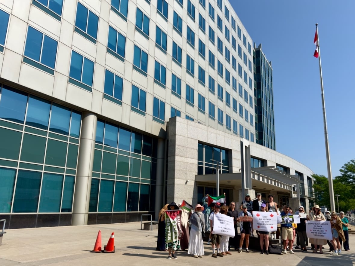 A group of people holding signs and flags pose together in front of an office building with a Canadian flagpole.