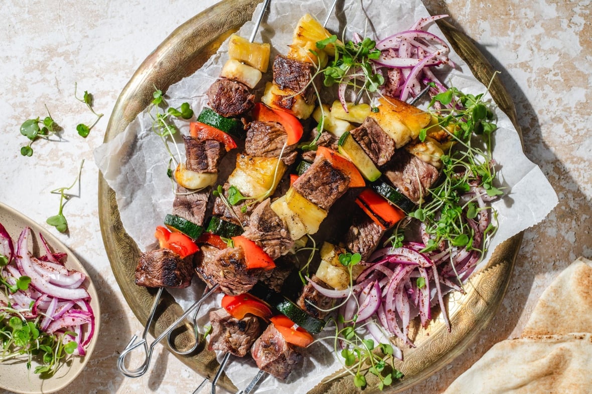 A platter of steak kebabs with pineapple and halloumi with a small red onion salad next to them.