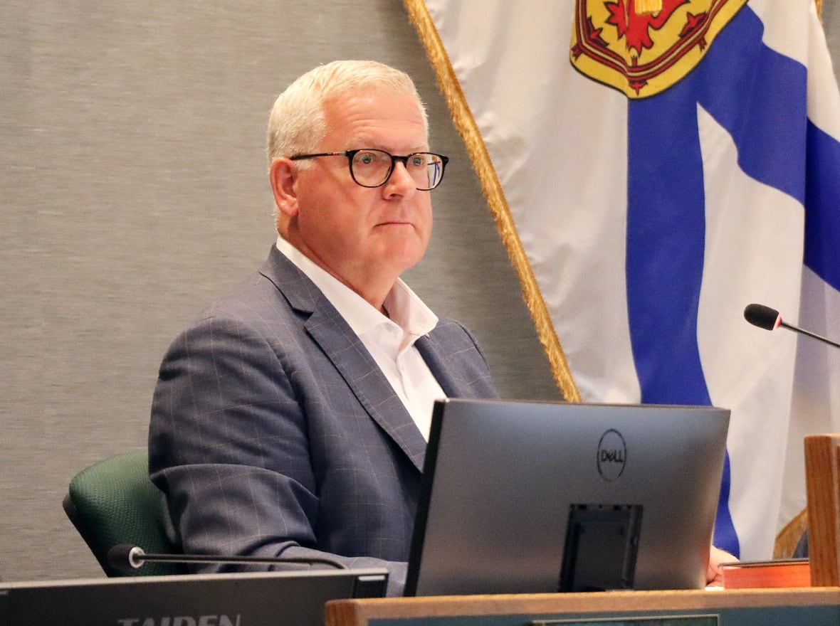 A man with white hair wearing glasses and a blue blazer with a white shirt open at the neck sits up straight behind a microphone with a Nova Scotia flag behind him.