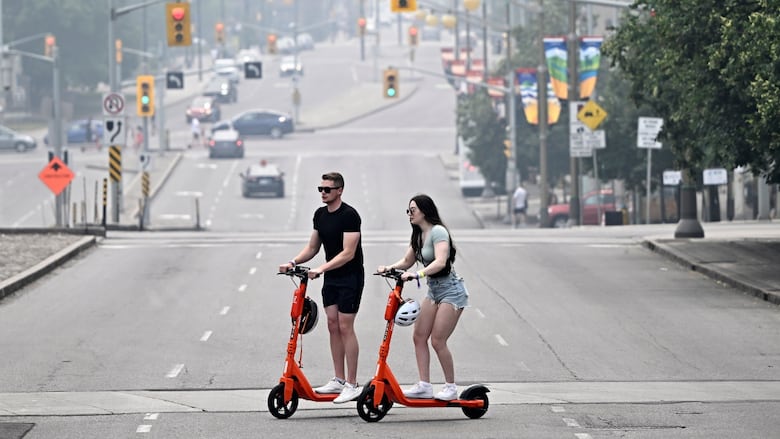 People cross a street on rental e-scooters with haze from wildfire smoke in Ottawa in June 2023.