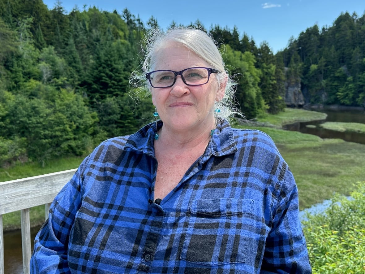 A woman looks into the distance, with forest and a watercourse in the background.