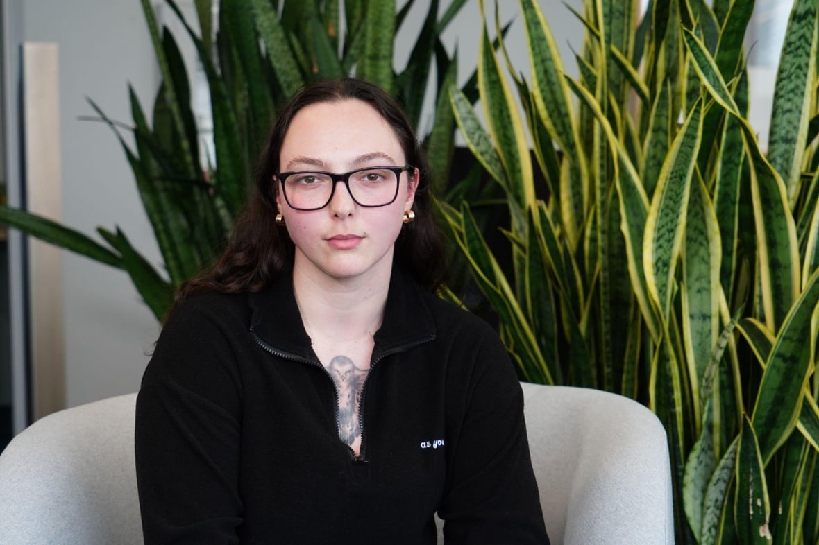 A woman looks at the camera. She is seated in front of a green plant.