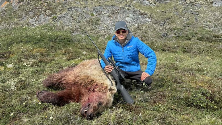 A man crouched next to a dead grizzly bear on a green tundra. 