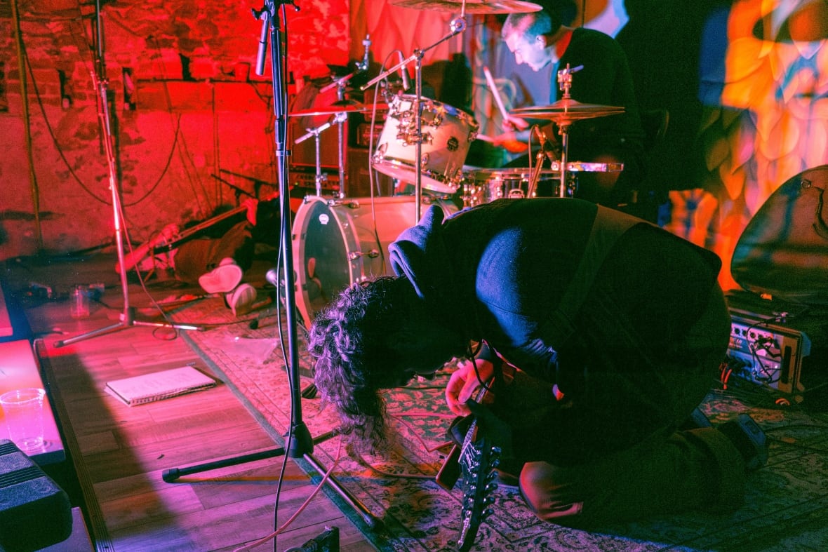 A crouching guitarist performing on a carpeted stage with pink lights.
