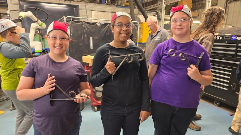 Three kids stand in a welding shop holding pot holder brackets.