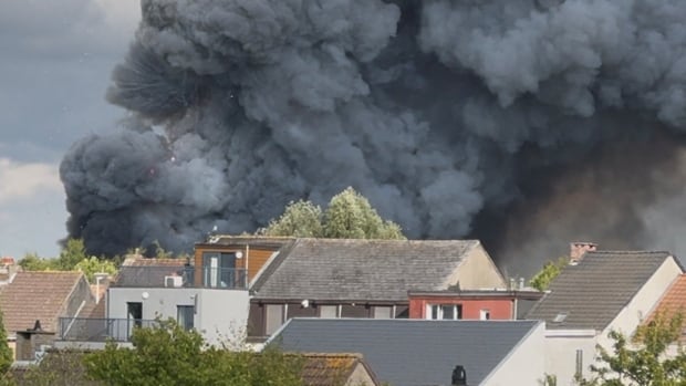 Fireworks burst and smoke rises as a fire engulfs the main stage at a music festival. Houses are in the foreground, but not burning.