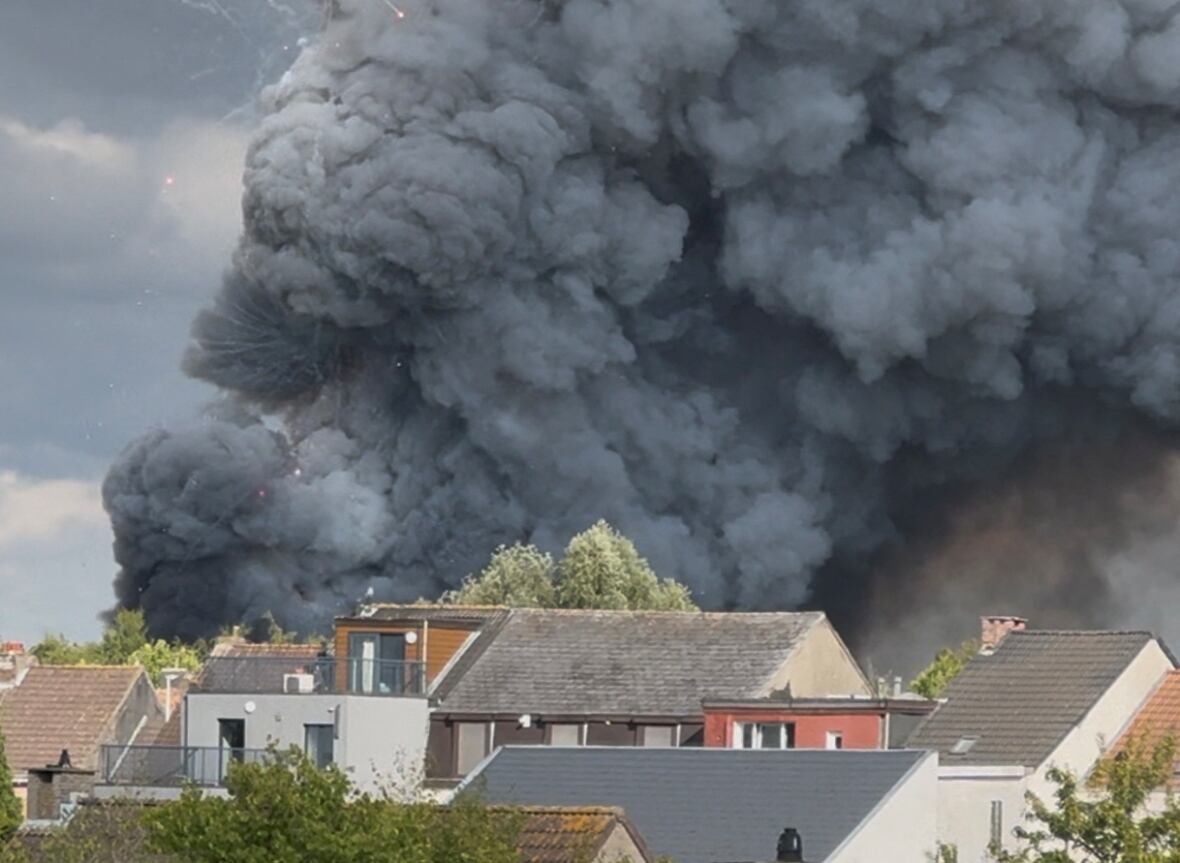 Fireworks burst and smoke rises as a fire engulfs the main stage at a music festival. Houses are in the foreground, but not burning.