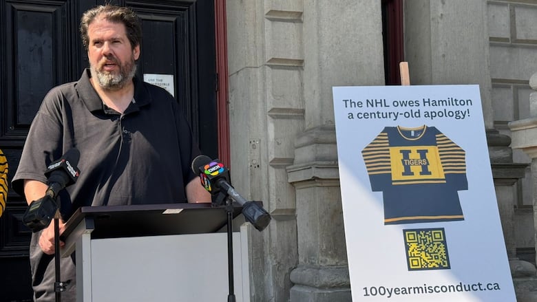 a tall person stands at a podium beside a poster saying the NHL owes Hamilton an apology