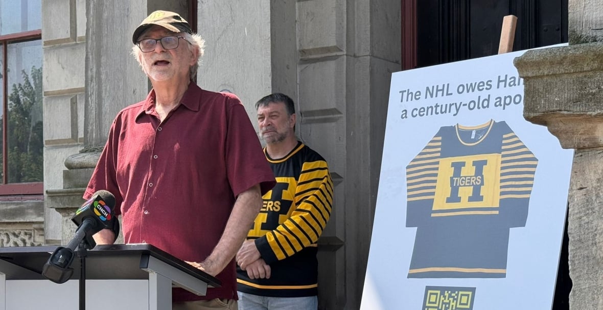Two peopel stand behind a podium. One is wearing a hockey jersey.