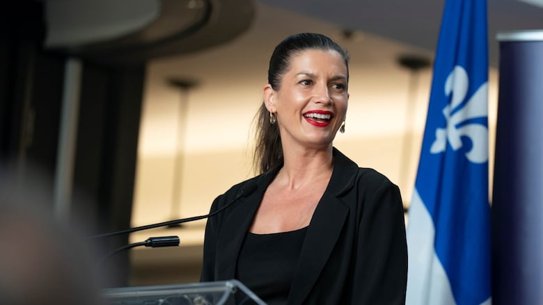 A woman with her hair in a slicked back pony tail smiles at a podium. The Quebec flag is behind her.