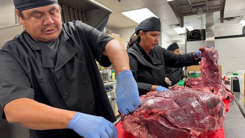 A man in the foreground and a woman and another person at the same counter, all wearing black food-preparation shirts, hats and gloves, cut meat in a commercial kitchen.
