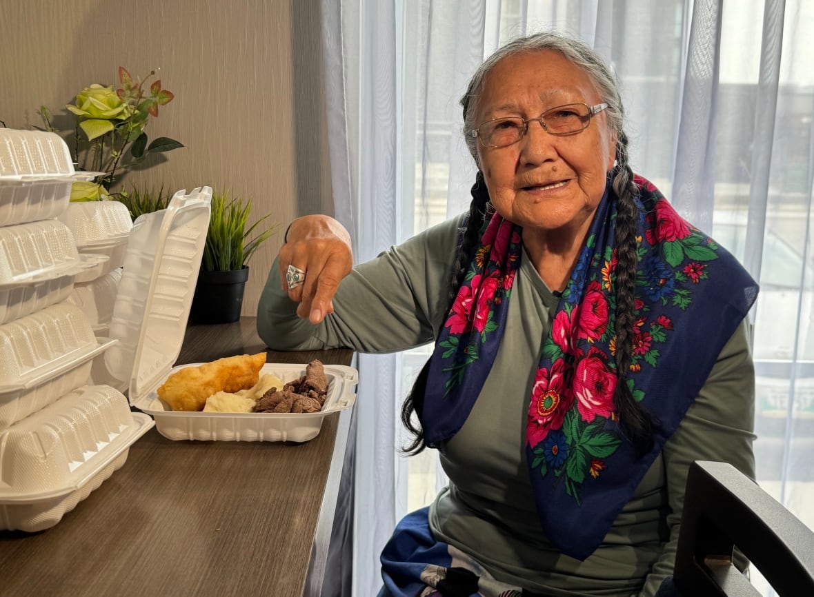 A woman with grey hair in braids smiles at the camera as her hand moves to the meat in a takeout container of food that also holds mashed potatoes and bannock.