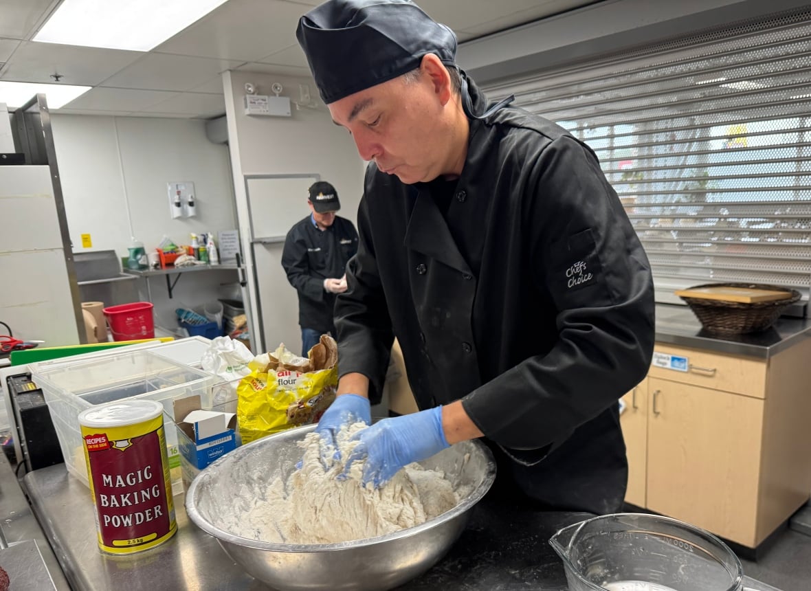 A person in food-preparation clothing works dough in a large metal bowl.