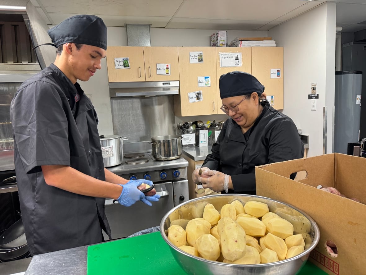 A man and a woman peel potatoes in a commercial kitchen. A large bowl of peeled potatoes sits on the counter in the foreground.
