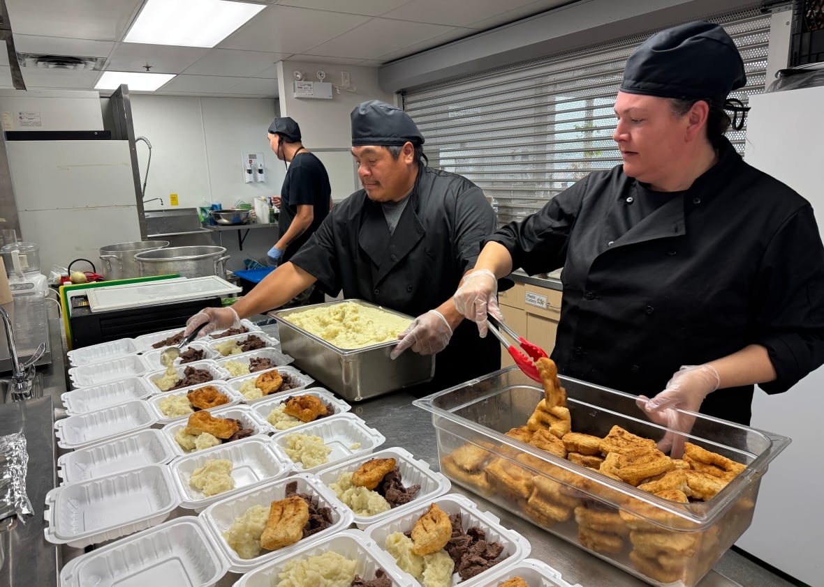 Two people add bannock and potatoes to takeout containers in a commercial kitchen.
