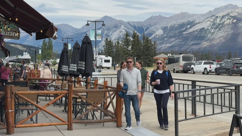 A few pedestrians walk on a sidewalk in Jasper with the mountains in the backdrop, on a cloudy day in July.