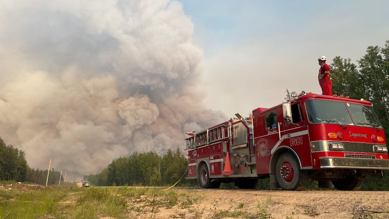 A firefighter stands on top of fire truck, looking out at large plume of smoke from a distant fire.